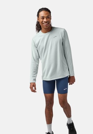 lichtgroen sportshirt met lange mouwen en een textuurpatroon, gecombineerd met marineblauwe compressiekorte broek en zwarte hardloopschoenen.