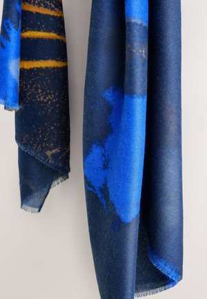 Dva visiace šály s abstraktnými vzormi v odtieňoch modrej a tmavomodrej, jeden so žltými horizontálnymi pruhmi a ošúchanými okrajmi.