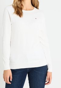 Valkoinen pitkähiha neulepusero pyöreällä kaula-aukolla ja hillityllä logolla rinnassa, yhdistetty tummansinisiin farkkuihin. Sileä pinta.