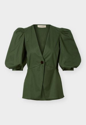 Mujer con top de manga abullonada verde oscuro y pantalones cortos a juego, gafas de sol negras, sandalias tejidas, sosteniendo un bolso de gran tamaño también tejido.