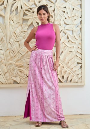 Strak lavendelkleurig tanktop gecombineerd met een lange, vloeiende roze rok met ingewikkelde patronen en een zijsplit, tegen een lichte, gestructureerde achtergrond.