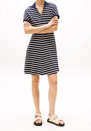 Persoon draagt een marineblauwe en wit gestreepte jurk met korte mouwen en kraag, samen met beige sandalen met open tenen, staand met gekruiste armen.