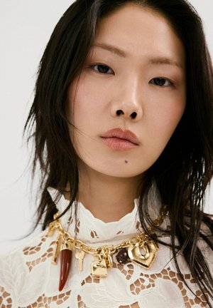 Joven con cabello lacio y oscuro que lleva una blusa blanca de encaje y un collar de cadena dorada con varios colgantes grandes, incluyendo un candado y una llave.