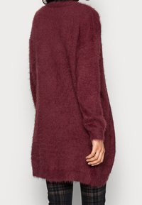 Lång, burgundy tröja gjord av mjukt, fluffigt material. Har en avslappnad passform, ribbad halsringning och något längre bak. Styler med rutiga byxor.