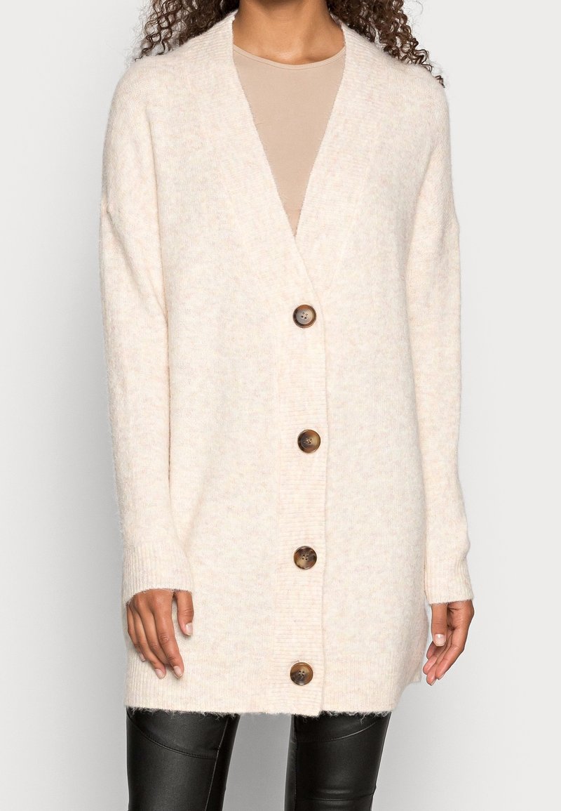 Vaaleanbeige neulejakku, valmistettu pehmeästä materiaalista, jossa on V-aukko, pitkät hihat ja viisi pyöreää ruskeaa nappia edessä.