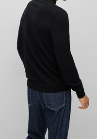 Svart stickad tröja med långa ärmar och en figurnära design, bärs över blå denimjeans med synliga sömmar och fickor.