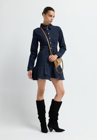 Femme portant une robe en denim foncé à boutons avec une jupe évasée, des bottes noires décontractées et un sac bandoulière beige, se tenant contre un fond blanc.