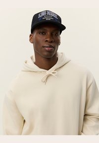 Crèmekleurige hoodie met een trekkoord en een zwarte pet met blauw geborduurde tekst "LES DEUX ATHLETIC DEPARTMENT." Zacht materiaal.