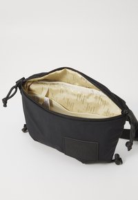 Black fabric bag with a zip closure, featuring a beige interior with logo patterns, adjustable straps, and a textured brand label on the front.