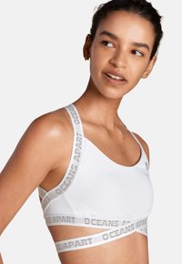 Weißer Sport-BH mit glatter Textur, ausgestattet mit einem breiten elastischen Bund mit "OCEANS APART" in Grau. Verstellbare Träger, minimalistisches Design.
