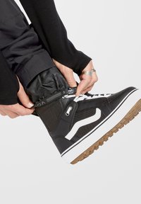 Zapatilla alta negra con raya lateral blanca, tejido texturizado, suela de goma y acentos metálicos, llevada mientras se cierra una prenda negra.