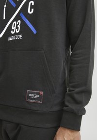 Black hoodie with a front pocket, featuring a graphic print in white and blue. Includes a label with "INDICODE" and a ribbed cuff.