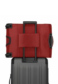 Borsa a duffle rossa in tessuto con accenti neri, manico retrattile e tasca in rete, poggiata su una valigia rigida grigia a nervature.