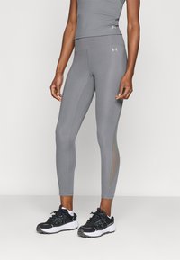 Sive sportske helanke s pripijenim krojem, kontrastnim svijetlim šavovima i umetcima od mrežaste tkanine sa strane. Uparene s crno-bijelim sportskim tenisicama.