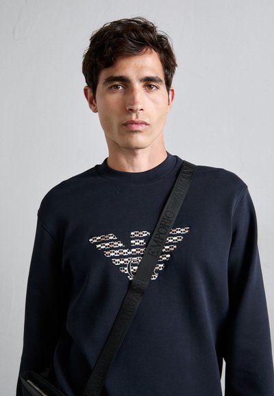Jongetje met donker haar draagt een marineblauwe sweatshirt met een patroon van een adelaarslogo en een zwarte schoudertasband met tekst.