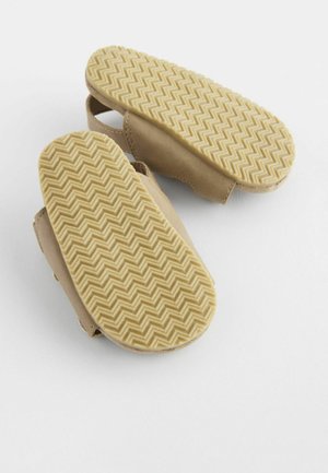 Zapatos suaves de color beige claro con suelas de goma que presentan un patrón en zigzag, abiertos en los laterales para facilitar su uso. Diseño ligero y flexible.