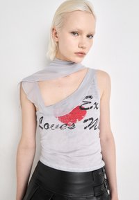 Débardeur gris clair avec un graphique de cercle rouge et du texte noir ; design asymétrique avec une superposition transparente à l'épaule. Jupe plissée noire.