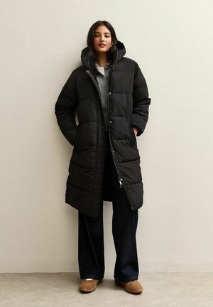 Abrigo acolchado negro con capucha, que cuenta con un cierre de botones y cremallera, patrón acolchado y bolsillos laterales, combinado con vaqueros de pierna ancha y zapatos tipo slip-on.