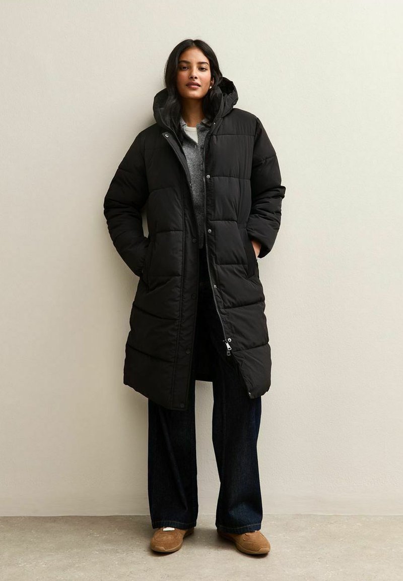 Cappotto piumino nero con cappuccio, dotato di chiusura con bottoni e zip, motivo trapuntato e tasche laterali, abbinato a jeans a gamba larga e scarpe slip-on.