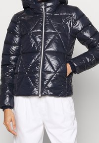 Mörkblå pufferjacka med blanka diamantquiltade mönster, framdragkedja, huva och elastiska ärmslut, tillverkad av glänsande syntetmaterial.