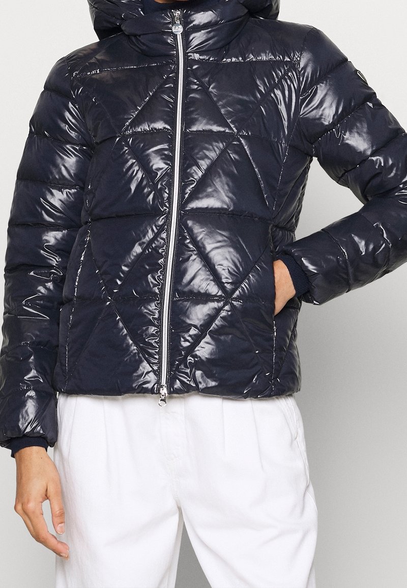 Mörkblå pufferjacka med blanka diamantquiltade mönster, framdragkedja, huva och elastiska ärmslut, tillverkad av glänsande syntetmaterial.