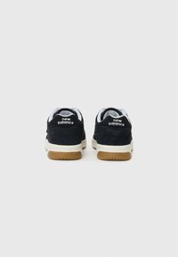 Crne suede tenisice s bijelim "new balance" natpisom na peti, kremastim srednjim dijelom i gumiranim potplatom. Kratko vezanje i podstavljeni ovratnik.