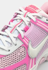 Nike Vomero 5 tenisice imaju bijelu podlogu s ružičastim naglasicama, teksturiranu gornjište s mrežastim panelima i bijele vezice. Brending se nalazi na jeziku.