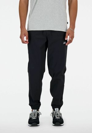 Mężczyzna w czarnych spodniach jogger New Balance, szarej koszulce i czarnych butach sportowych New Balance, stojący na białym tle.