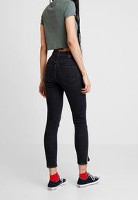 Pantalones vaqueros negros ajustados con un diseño ceñido, que cuentan con bolsillos traseros y un ligero deshilachado en el tobillo, combinados con una camiseta corta verde y zapatillas.