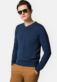 Granatowy sweter w serek z miękkiego dzianiny, z subtelnym logo na klatce piersiowej oraz ściągaczami przy mankietach i u dołu. Noszony na beżowych chinosach.