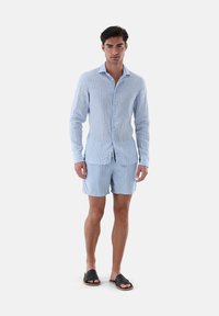Helles blaues gestreiftes Hemd und Shorts-Set. Das Hemd hat lange Ärmel und eine Knopfverschluss; die Shorts enden oberhalb des Knies. Getragen mit schwarzen Sandalen.