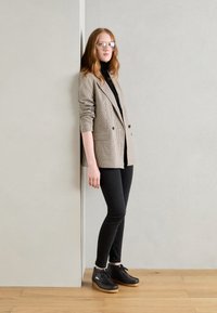 Beige és fekete houndstooth mintás blézer, fekete garbóval, skinny fekete farmernadrággal és barna talpú fekete cipővel, egy szürke fal előtt.