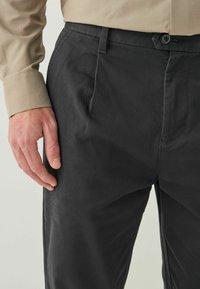 Čierne nohavice s hladkým povrchom, slim fit, so bočnými vreckami a zapínaním na gombík, v kombinácii s béžovou košeľou s dlhým rukávom.