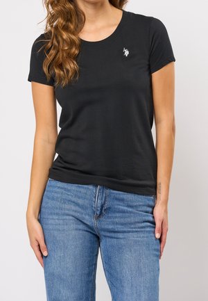 Femme portant un t-shirt noir uni à manches courtes avec un petit logo blanc de joueur de polo sur la poitrine et un jean bleu, debout devant un fond clair.