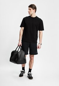 Svart bomull T-skjorte, svarte shorts, holder en svart lær treningsbag. Mørke sneakers med hvite detaljer, minimalistisk design og rene linjer.