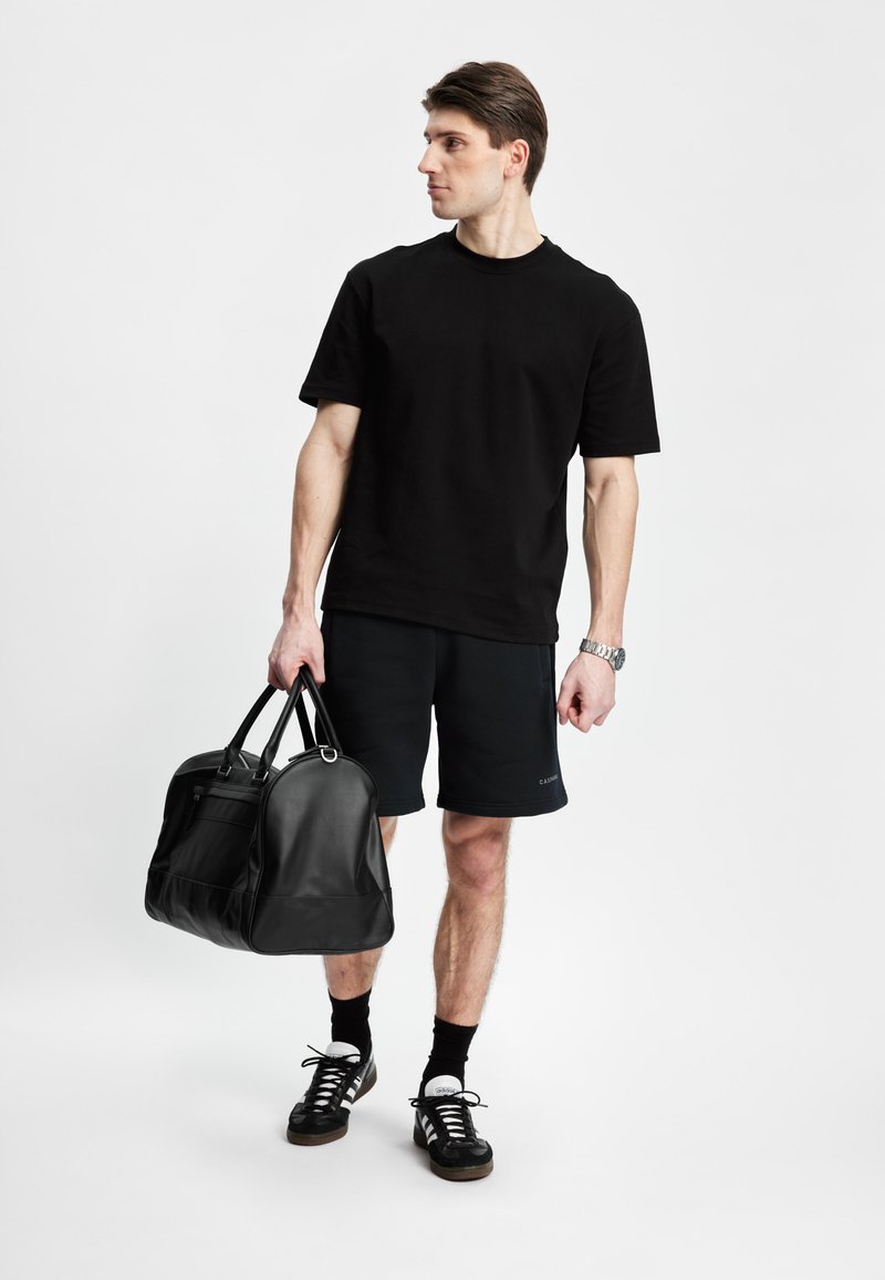 Svart bomull T-skjorte, svarte shorts, holder en svart lær treningsbag. Mørke sneakers med hvite detaljer, minimalistisk design og rene linjer.