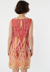 Robe sans manches dans des teintes vives de rouge et d'orange avec des motifs géométriques. Présente une découpe en forme de clé au dos. Tissu doux et léger.