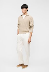 Άνδρας που φοράει μπεζ πλεκτό πουλόβερ, λευκό παντελόνι και καφέ σουέντ αθλητικά παπούτσια, στέκεται μπροστά σε απλό λευκό φόντο.