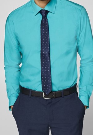 Mężczyzna ubrany w turkusową koszulę zapinaną na guziki, granatowy krawat w kropki, granatowe spodnie i czarny pasek, z rękami w kieszeniach.