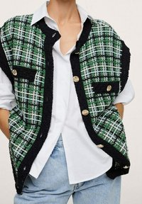 Groen, wit en zwart geblokt vest met gouden knopen, gedragen over een wit shirt en lichtblauwe jeans, handen in de zakken.