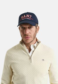 Marinepet met "GANT" in het rood geborduurd, met een gebogen rand. Crème-kleurige, gestructureerde ritstrui gedragen over een wit shirt.
