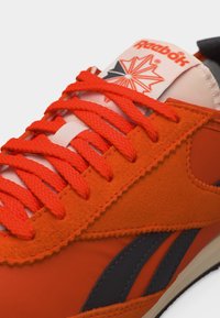 Πορτοκαλί αθλητικό παπούτσι Reebok με υφασμένα πάνελ από σουέντ, φωτεινά πορτοκαλί κορδόνια και λογότυπο στη λευκή γλώσσα με κόκκινο και μαύρο σχέδιο.