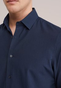 Man draagt een marineblauw overhemd met knopen en een puntige kraag, met zicht op de bovenkant van de borst en het onderste deel van het gezicht tegen een neutrale achtergrond.