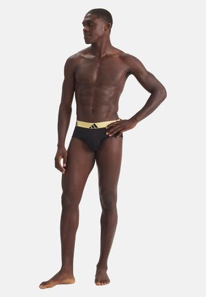 Hombre atlético de piel oscura de pie descalzo, vistiendo un bañador negro con una amplia cintura amarilla que presenta el logo negro de Adidas.