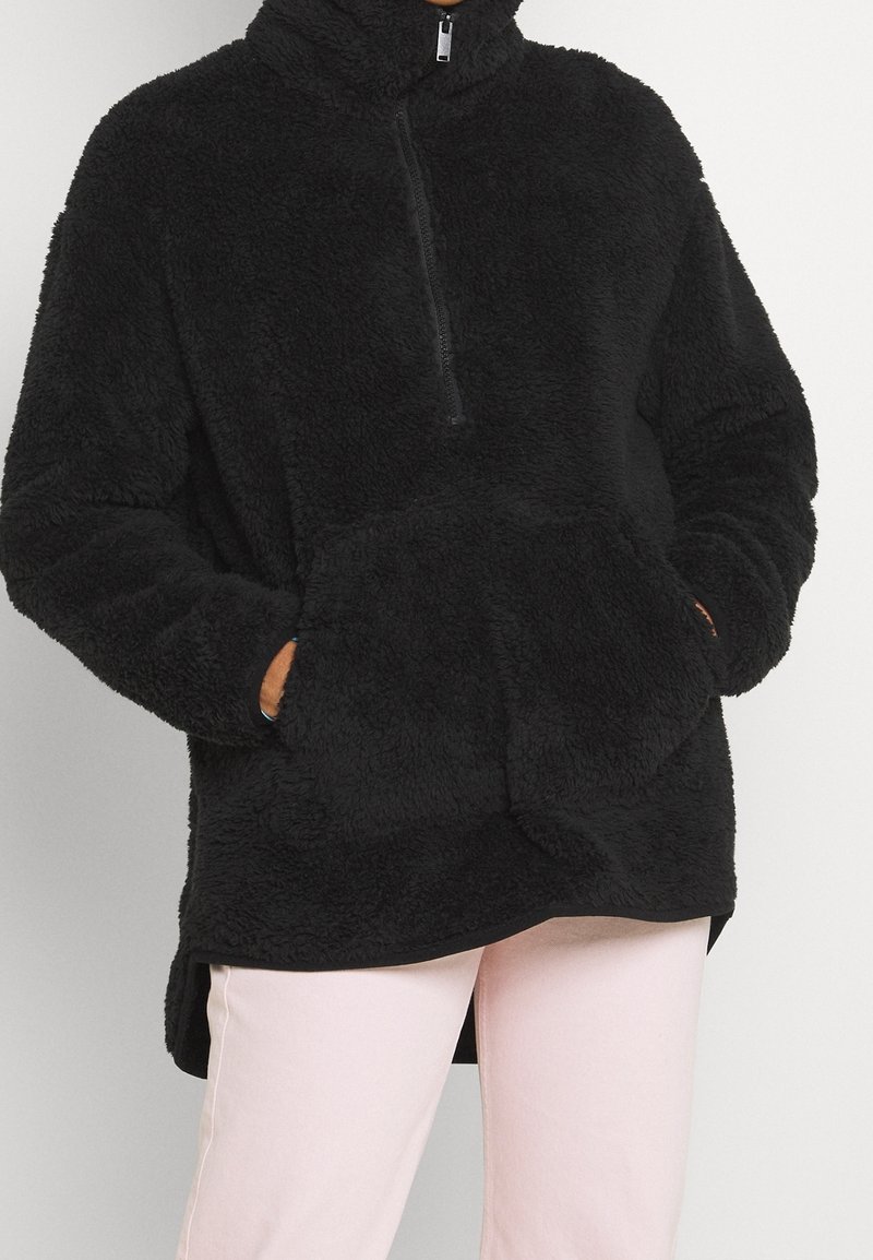 Personne portant un pull noir en tissu duveteux avec une fermeture éclair frontale et une poche kangourou, assorti à un pantalon rose clair, les mains dans les poches.