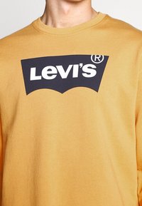 T-shirt gialla a maniche lunghe in cotone, con un logo Levi's nero in un design curvo sul petto. Stile semplice e casual.