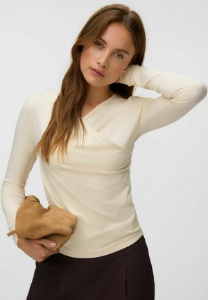Jeune femme aux longs cheveux bruns portant un haut à manches longues crème et un pantalon noir, tenant un article en daim marron plié dans une main.