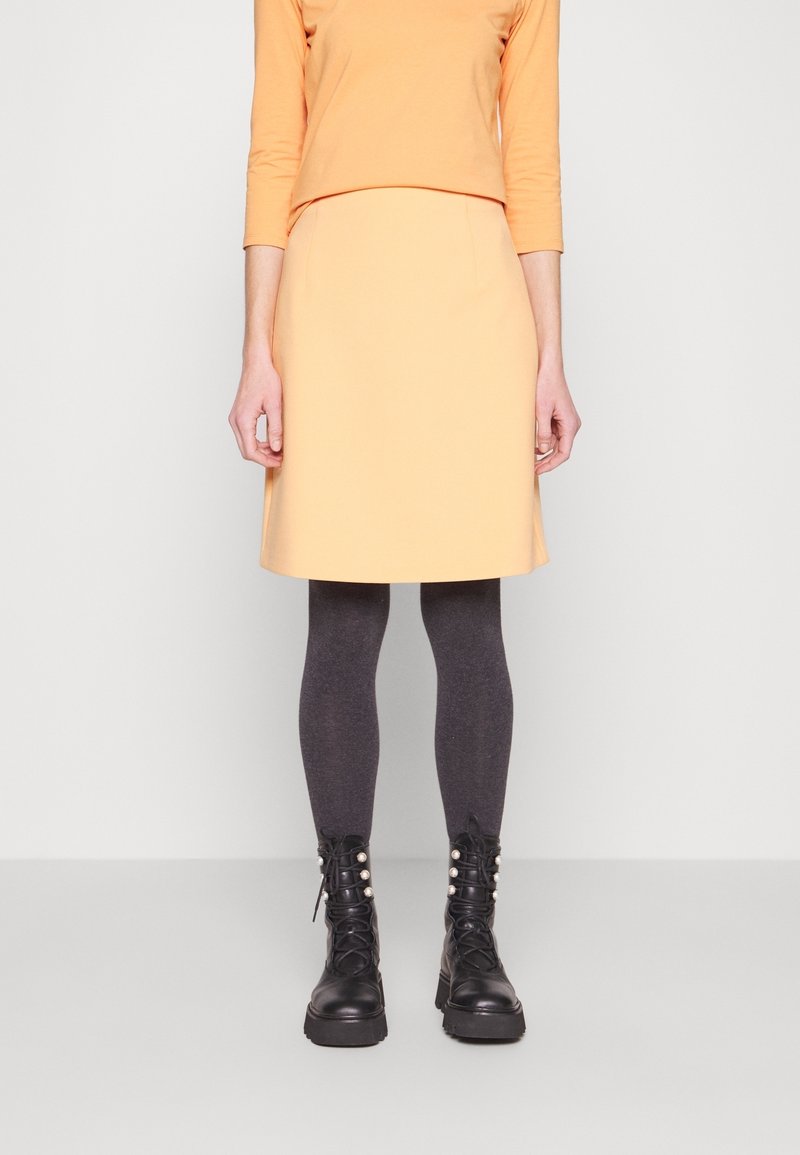 Femme portant un haut à manches longues pêche et une jupe, des collants gris foncé et des bottes noires à lacets avec des ornements en perles.