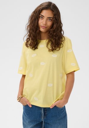 Ung kvinde med krøllet hår iført en gul t-shirt med hvide ovale mønstre og blå jeans, hænderne i lommerne, på en hvid baggrund.