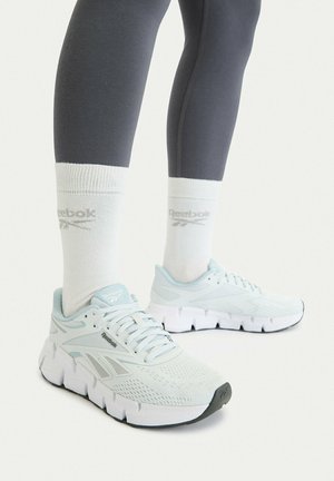 Hellblaue Reebok Laufschuhe, getragen mit weißen Reebok-Socken und dunkelgrauen Leggings vor weißem Hintergrund.